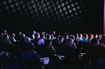 Audience of IT executives in a dark theater viewing a keynote presentation and taking notes 