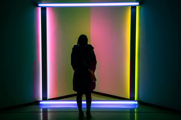 Silhouetted person standing in front of a vibrant neon light installation with vertical and horizontal tubes glowing in blue, pink, and yellow